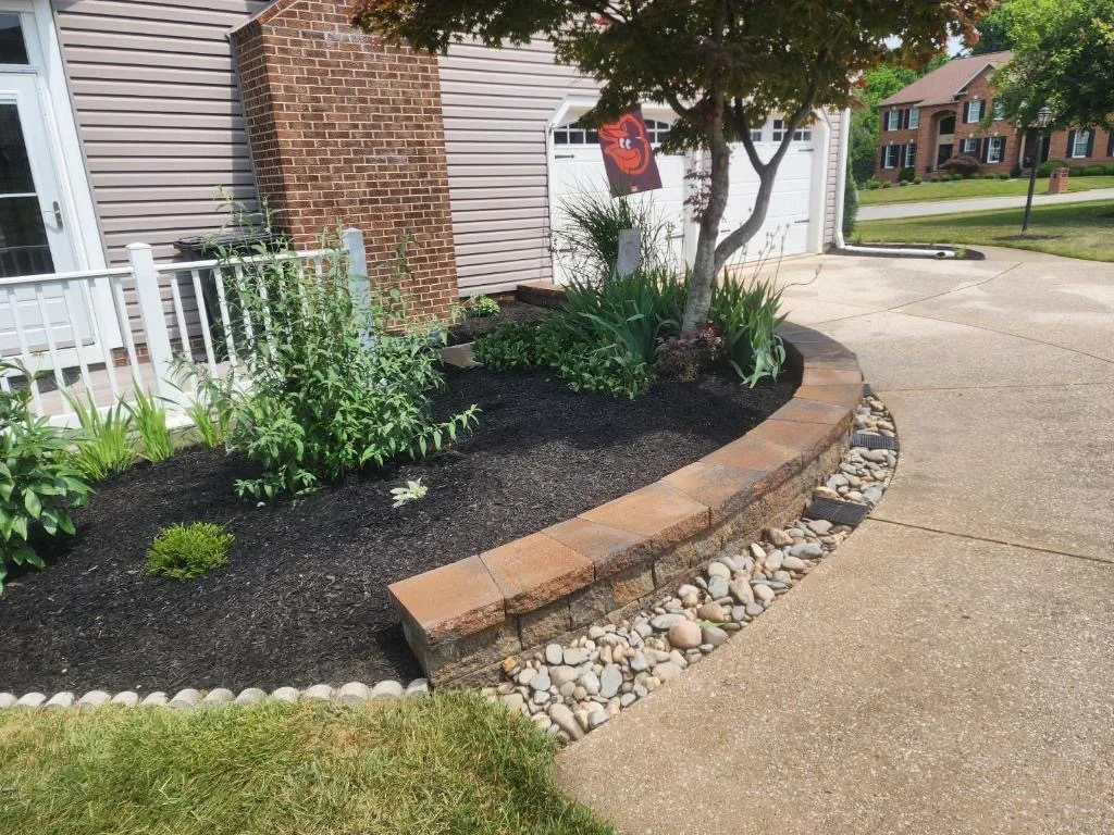 Curved landscape bed with black mulch, plants, and river rock edging along driveway