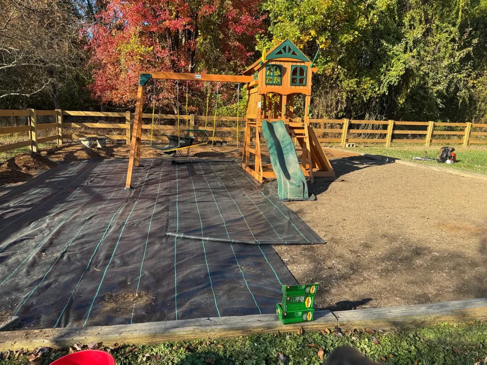 Black erosion control fabric with green rope anchoring on wooden stakes along streambank with wooden fencing