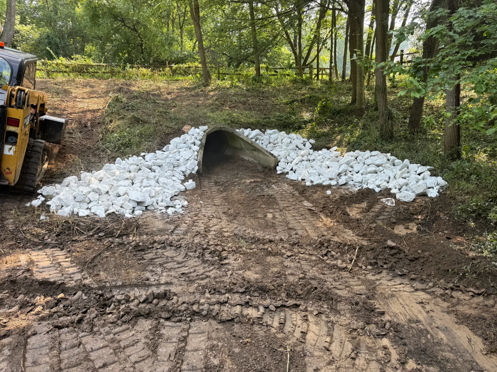 Concrete culvert pipe with white riprap stone stabilization in wooded area, equipment visible