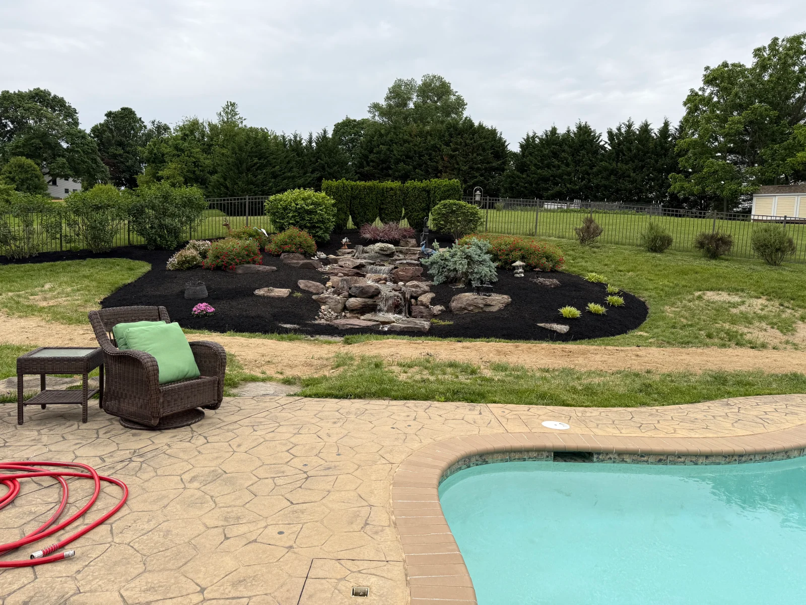 Black mulch landscape bed with stacked stone water feature, shrubs, and evergreens beside pool patio